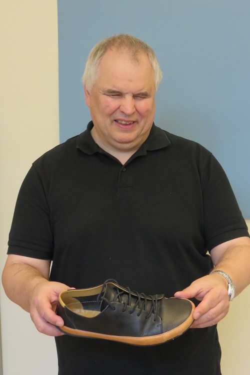 Bruno Kühne mit einem Barfußschuhmodell, das er selbst trägt Bruno Kühne hält lächelnd einen grauen Barfußschuh in beiden Händen.
