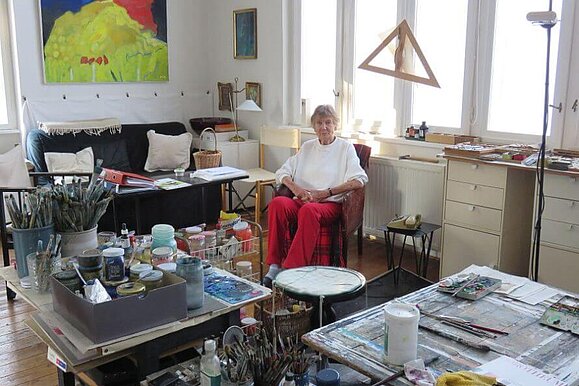 Lucia Kellner im Atelier ihres Hauses Eine Frau sitzt in einem Raum mit vielen Malutensilien