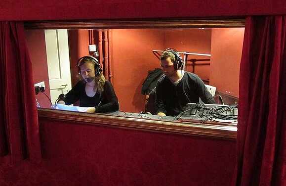 Kommentatorin Stefanie Panzenböck und ein Tontechniker bei der Arbeit im Burgtheater. (c) AUDIO2 Frau und Mann mit Kopfhörern in roter Theaterloge.