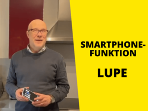 Daniele zeigt die Smartphonefunktion Lupe. Mann mit grauem Pullover steht in Küche mit Smartphone, rechts Text auf gelbem Hintergrund: "Smartphonefunktion Lupe"