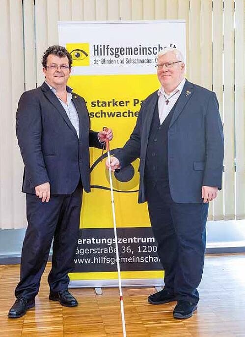 Zwei Männer mit Anzug vor Hilfsgemeinschaft Roll-Up, halten beide weißen Stock