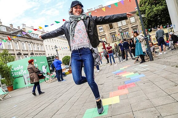 Eröffnung der "Coolen Straße" am Karmeliterplatz im Juni, Copyright: Mobilitätsagentur Wien/Christian Fürthner Junge Frau in Jeans tanzt mit ausgebreiteten Armen über die bunten Felder der "Coolen Straße" am Karmeliterplatz.
