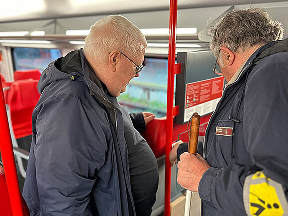 Zwei Männer stehen in einem modernen Zug vor einem Knopf. Beide tragen Jacken, einer hält einen Taststock und trägt eine gelbe Armbinde. Er drückt den Knopf, während ihn beide betrachten. 