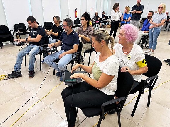 Eine Gruppe von Menschen sitzt in einem hellen Raum auf Stühlen, einige von ihnen halten elektronische Geräte mit Kabeln. Sie wirken konzentriert und haben Spaß, während sie an einer interaktiven Aktivität teilnehmen. Im Hintergrund stehen weitere Personen, die das Geschehen beobachten. Eine Frau trägt eine gelbe Armbinde und einen gelben Button mit drei schwarzen Punkten.