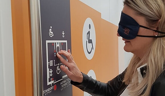 Eine Frau mit Augenbinde ertastet einen taktilen weißen Orientierungsplan, der mit dunkler Farbe hinterlegt ist.