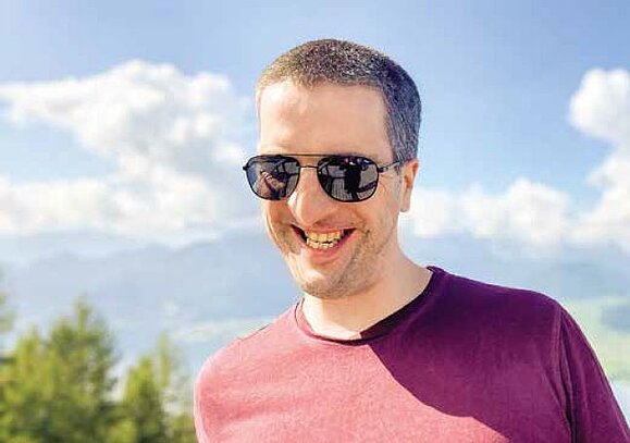 Blind_unterwegs_Fredrik_Fischer_Hilfsgemeinschaft.JPG lachender Mann mit Sonnenbrille und purpur Shirt vor blauem Himmel