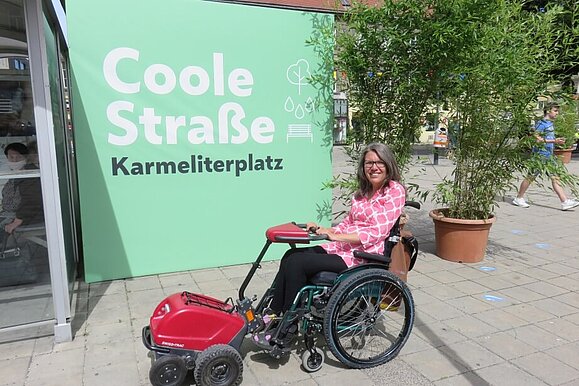 Maria Grundner ist mit der Barrierefreiheit der Coolen Straßen zufrieden. Maria Grundner sitzt im Rollstuhl vor der grünen Plakatwand der "Coolen Straße" am Karmeliterplatz.