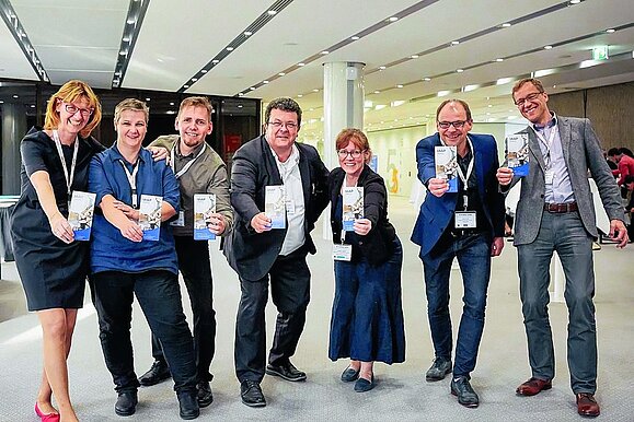 7 Menschen in Businesskleidung und mit Umhängeschild strecken Flyer mit Aufschrift "IAAP" in die Kamera.